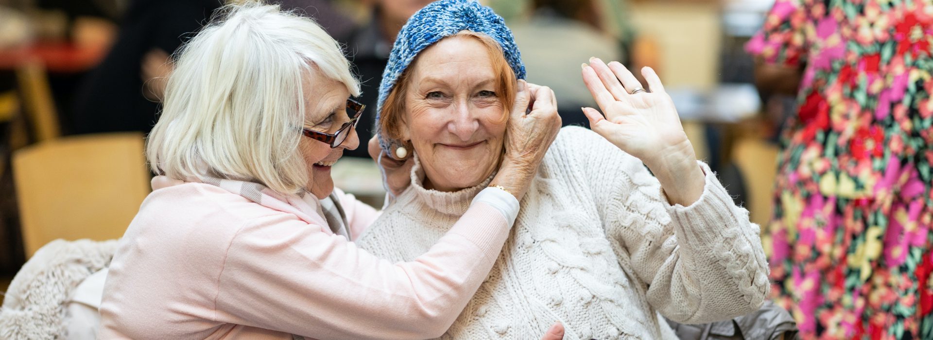 Two old white women who look like they are friends. The woman on the right handside is smiling and waving at the camera, the women on the lefthand side is fixing her friend's had. 