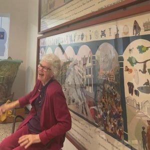 Sheila Dawkins sitting in front of the Bristol Tapestry at Bristol Museum and Art Gallery. Sheila is laughing.