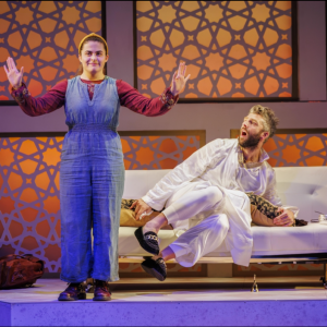 A still of the Arabian Nights theatre play at the Bristol Old Vic showing a woman dressed in blue smiling and holding her hands up in the foreground and a man dressing in white lying on a chaise longue in the background.