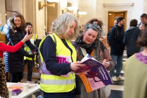 Two women standing in city hall and viewing a copy of International Women's Day programme