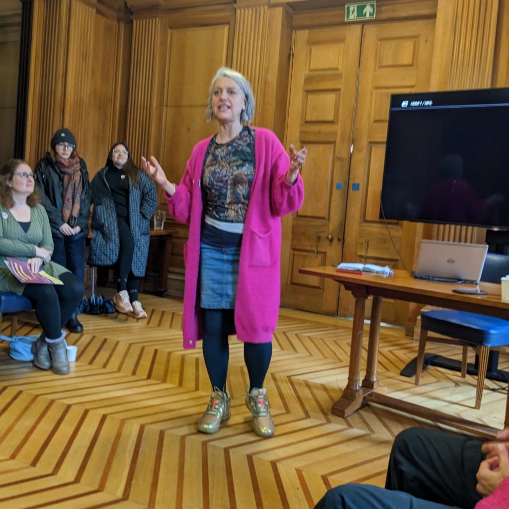 A woman in a bright pink cardigan standing in a room and talking to an audience at International Women's s Day 2024