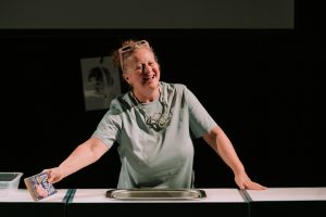 A woman on a theatre stage, smiling and holding a book called Flour Babies in her right hand.